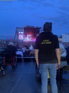 Photograph captures an outdoor concert by The Cure. Amanda Drollinger, wearing a black “AUDIO DESCRIBER” gilet stands on an accessible audience area. She and a wheelchair user, face a crowded stage, lit red and pink, with large video screens, metal staging. A blue evening sky shows fantastic weather, appreciated by very full crowd.